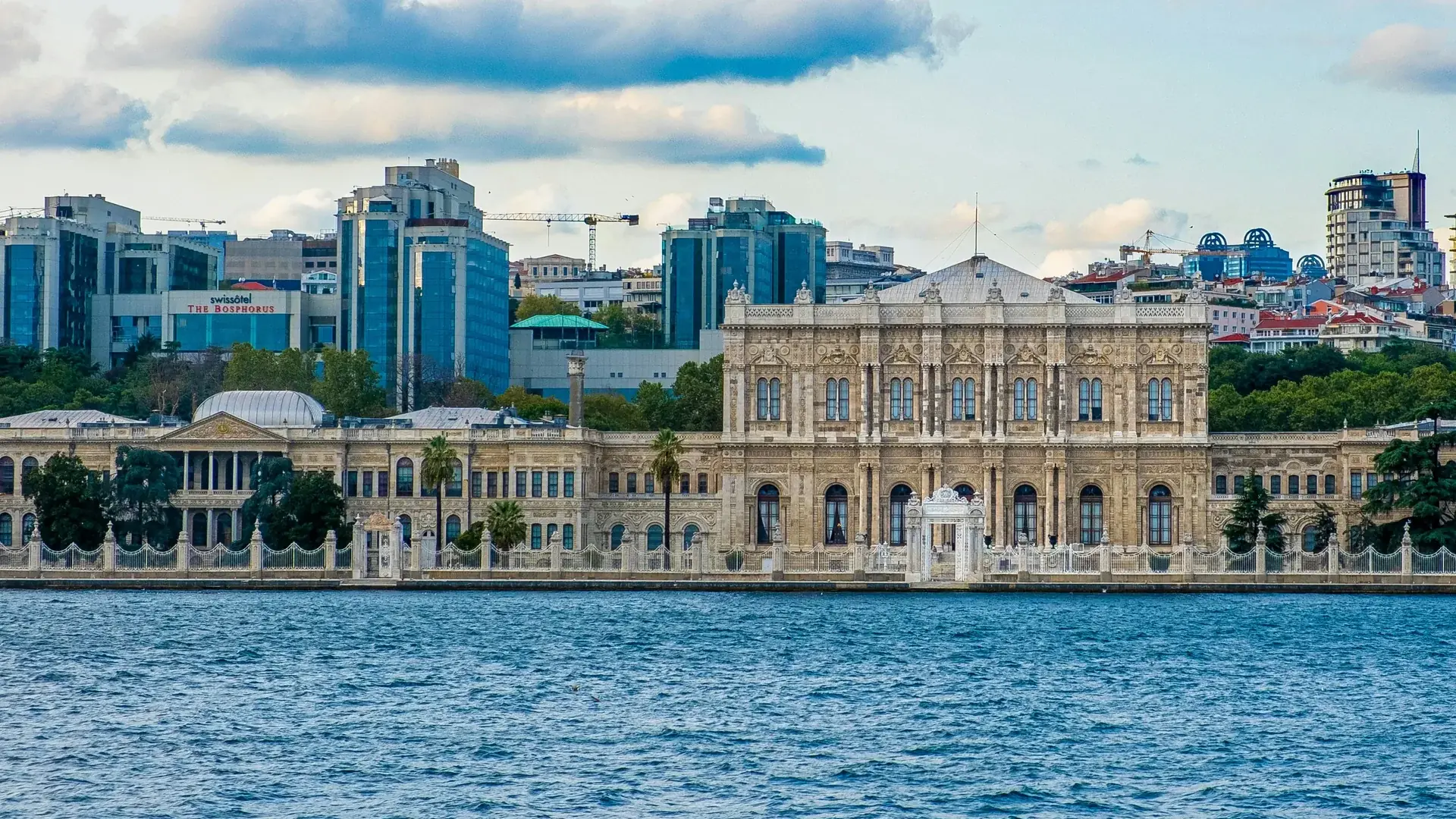 ボスポラス海峡越しに望むDolmabahçe Palace：中央棟を備えた長い大理石のファサード、岸壁の白い門、そして目の前に広がる青い海。